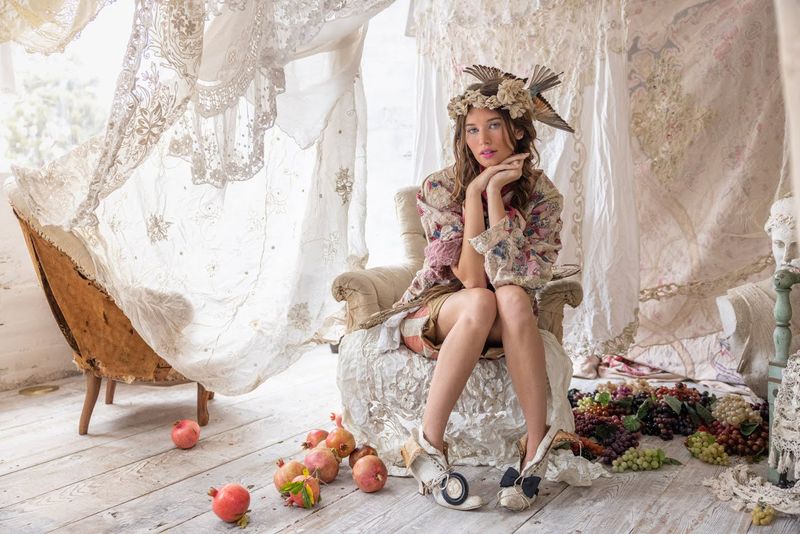 Woman in Magnolia Pearl vintage artisan clothing and whimsical hat, seated among lace, fruit, rustic decor.