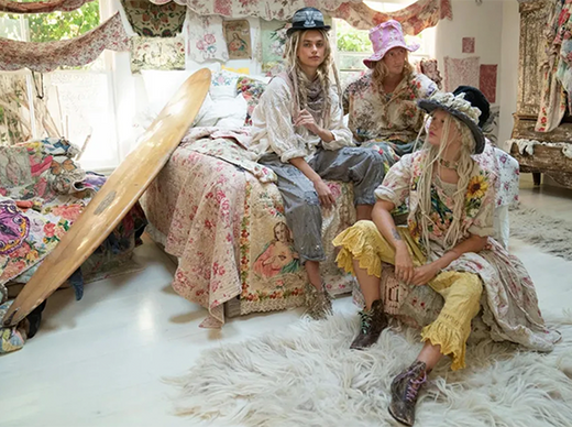Three women in whimsical Magnolia Pearl artisan clothing with vintage style, seated in a bohemian bedroom with floral bedding and eclectic decor.