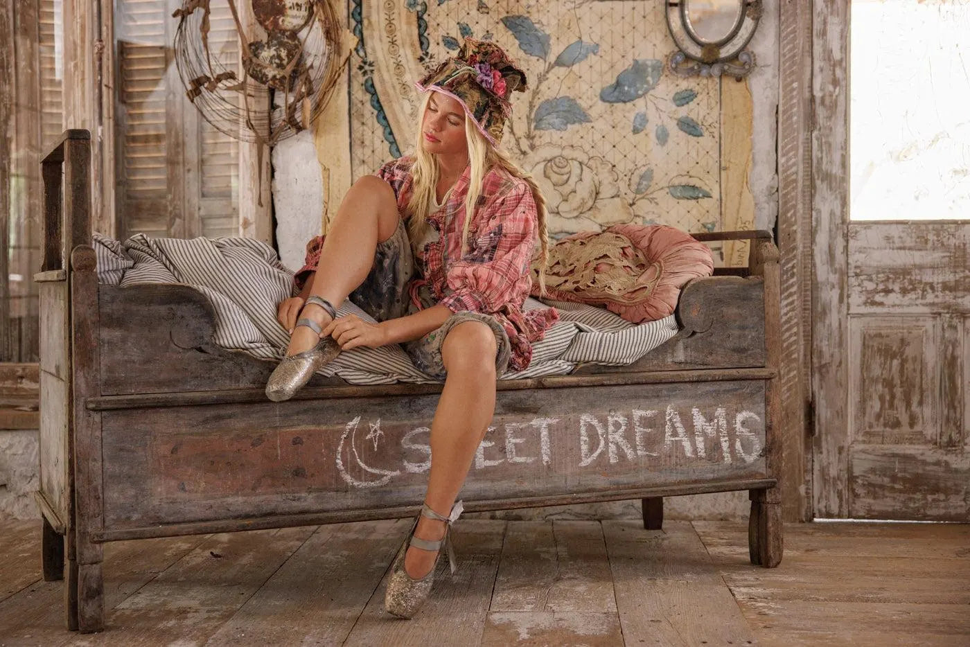Woman in vintage floral hat sitting on rustic bed with 'SWEET DREAMS' chalk sign in cozy room
