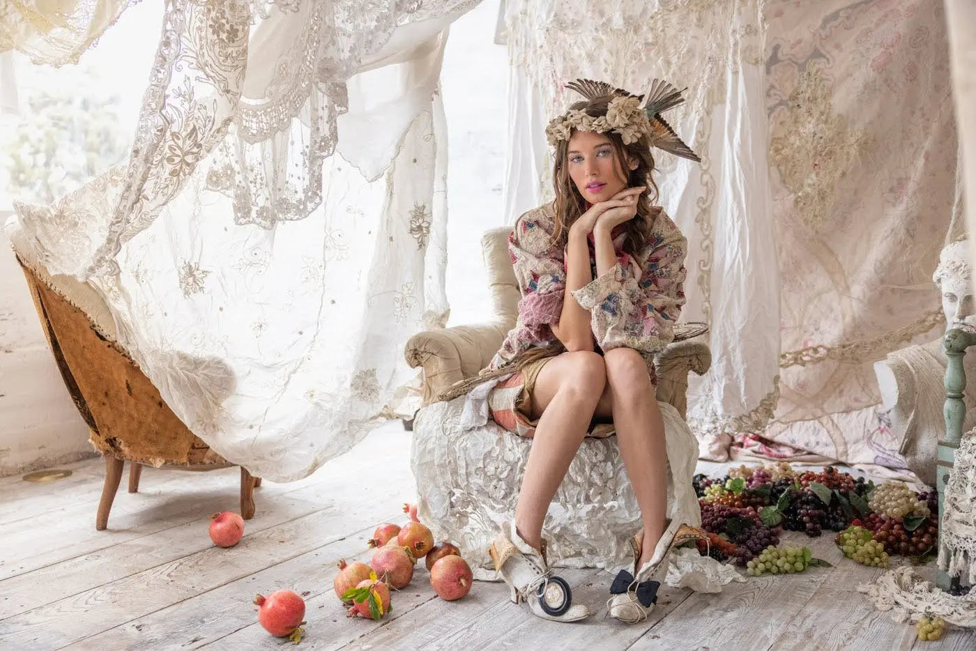 Woman in Magnolia Pearl artisan vintage clothing and whimsical headpiece, seated in lace-filled rustic room with pomegranates and grapes on the floor