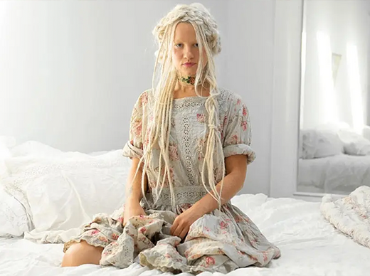 Woman in Magnolia Pearl artisan dress with whimsical vintage floral design sitting on white bed