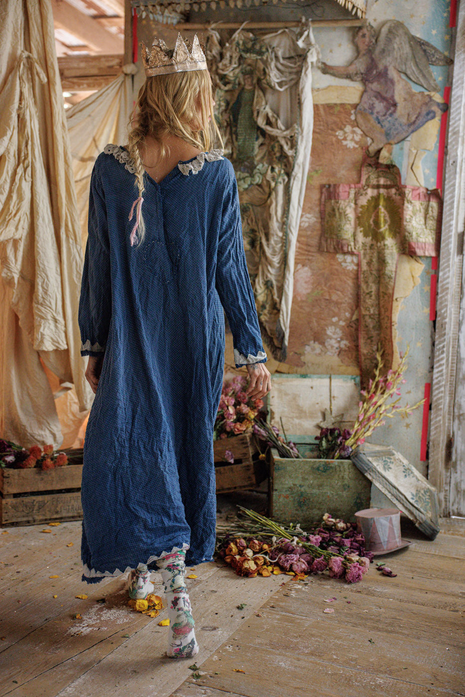 Woman in blue vintage Magnolia Pearl dress, lace trim, paper crown, artisan whimsical style, floral socks, surrounded by dried flowers in rustic room