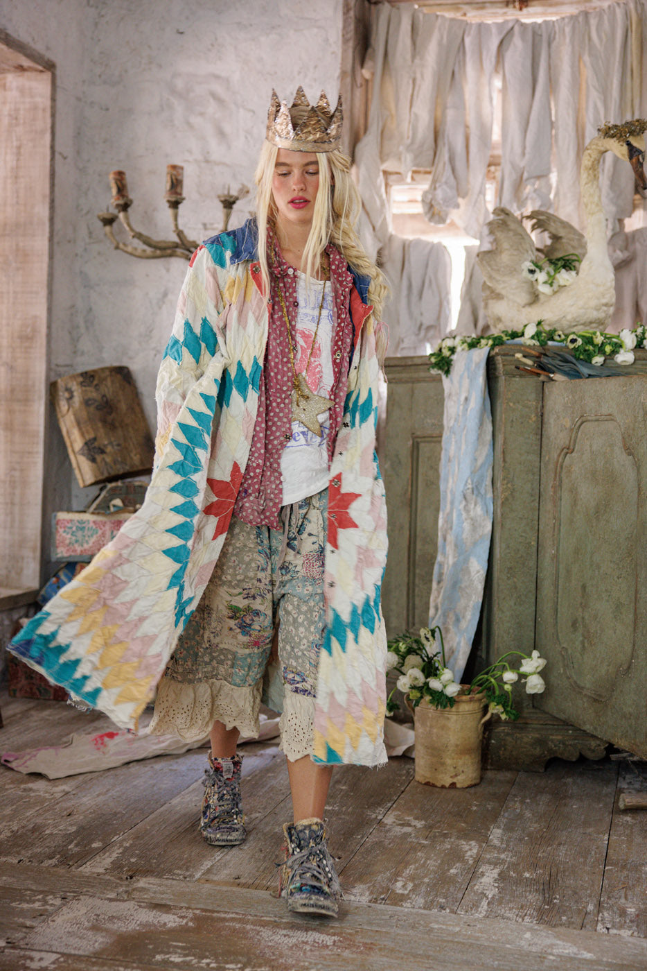 Woman in a patchwork quilt coat and crown standing in a rustic room with floral decor.