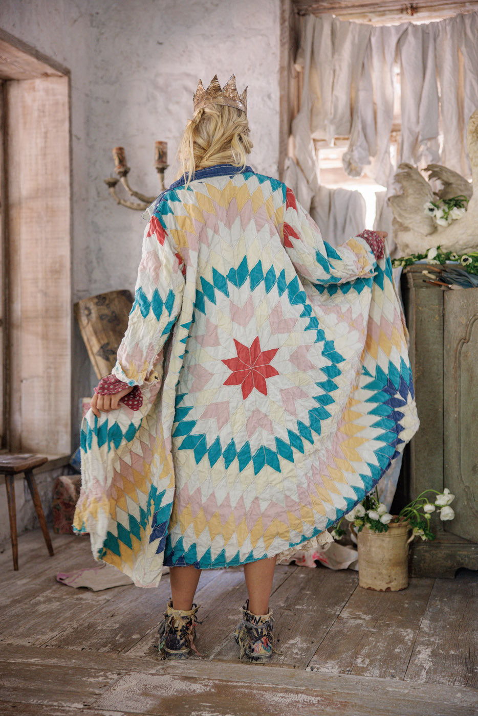 Woman in a colorful patchwork quilt jacket and crown stands in a rustic vintage room