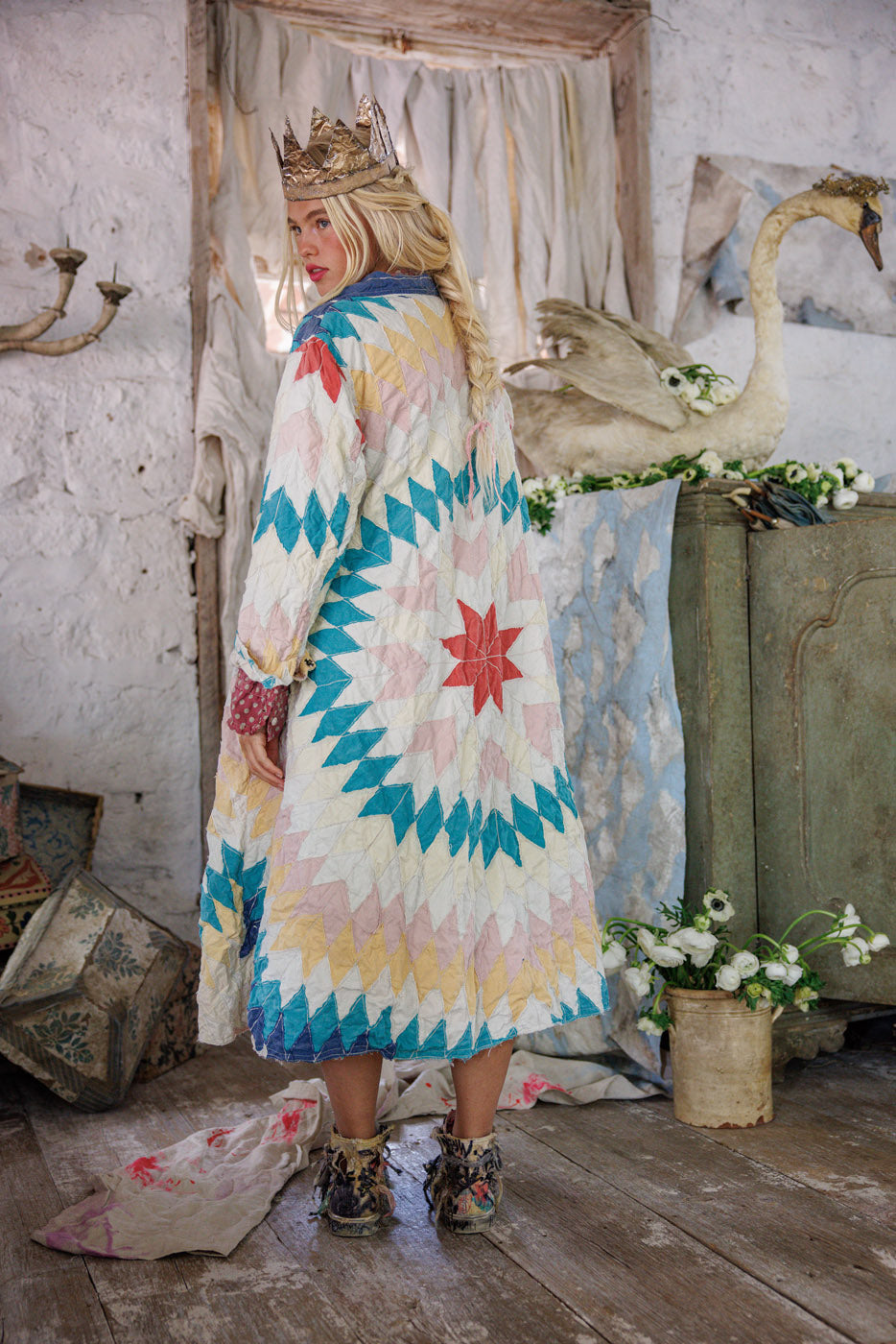 Woman in a patchwork quilt coat and crown standing indoors, rustic decor, floral accents