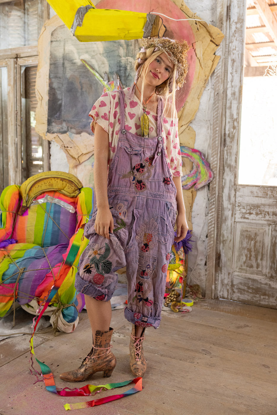 Person wearing a artistic overalls with ladybugs and floral embroidery and applique in a colorful, artistic indoor setting.