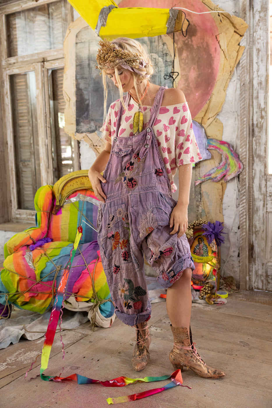 Woman in a colorful, eclectic setting wearing a heart t-shirt and purple ladybug overalls.