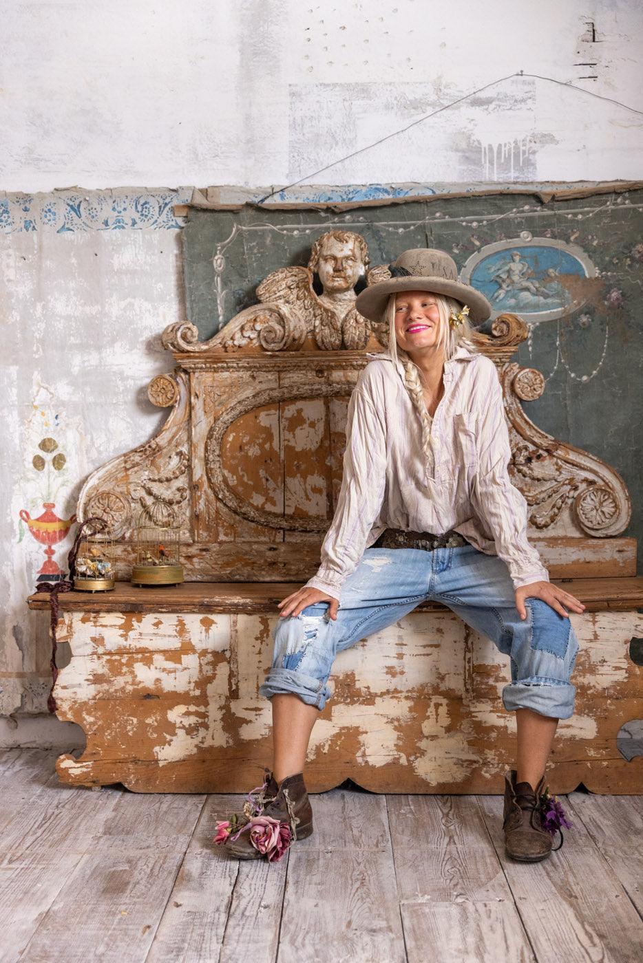 Woman in Magnolia Pearl artisan vintage shirt, hat, and jeans, sitting on distressed bench in whimsical rustic setting