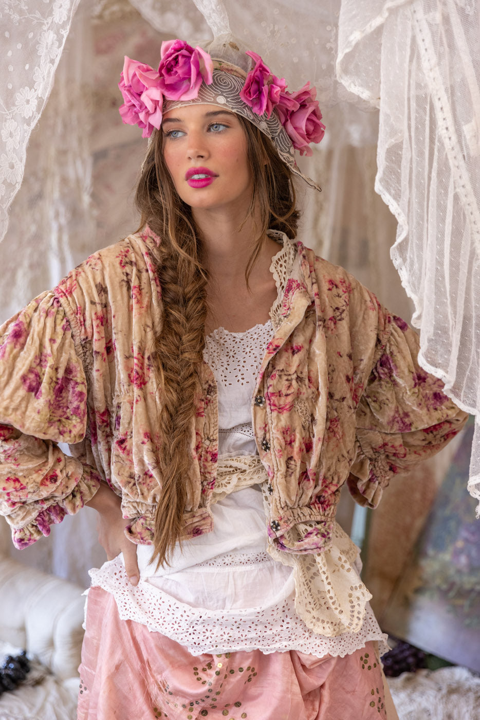 Model in Magnolia Pearl artisan vintage clothing, whimsical floral jacket, lace, and pink skirt, boho style, surrounded by lace curtains and grapes
