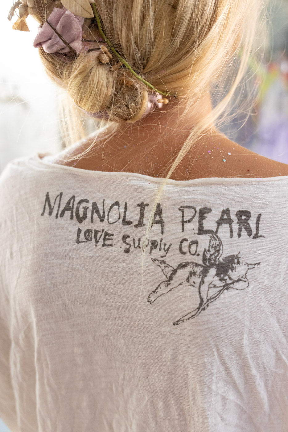 Back view of a person wearing a white t-shirt with 'Magnolia Pearl Love Supply Co.' text and logo.